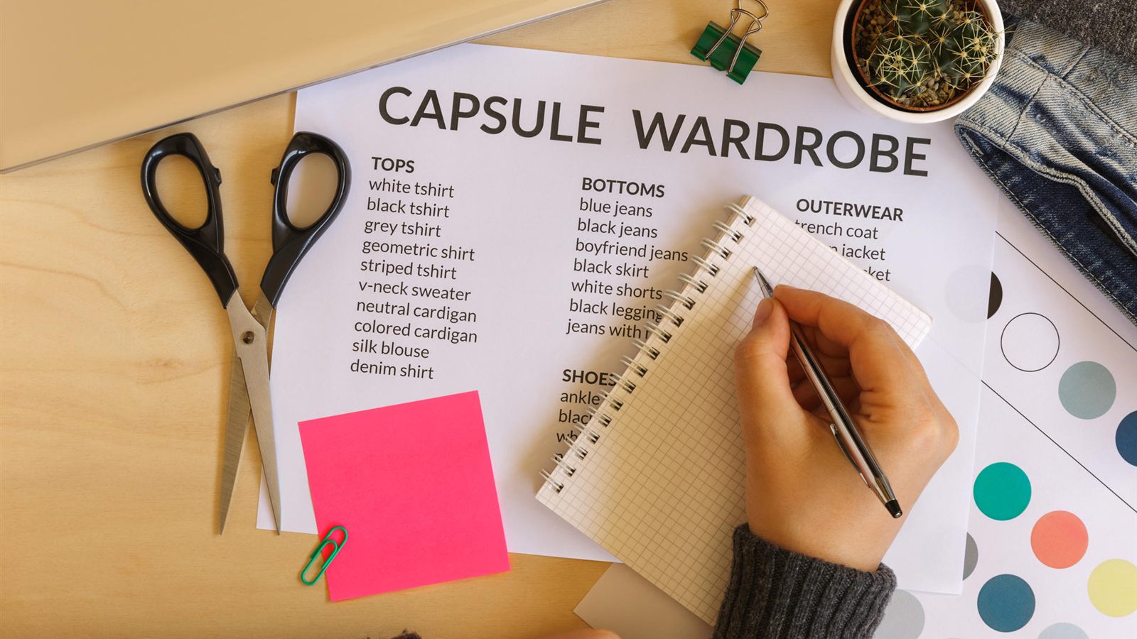 A hand writing in a notebook on top of a printed capsule wardrobe checklist of tops, bottoms, outerwear and shoes, next to scissors and colour swatches
