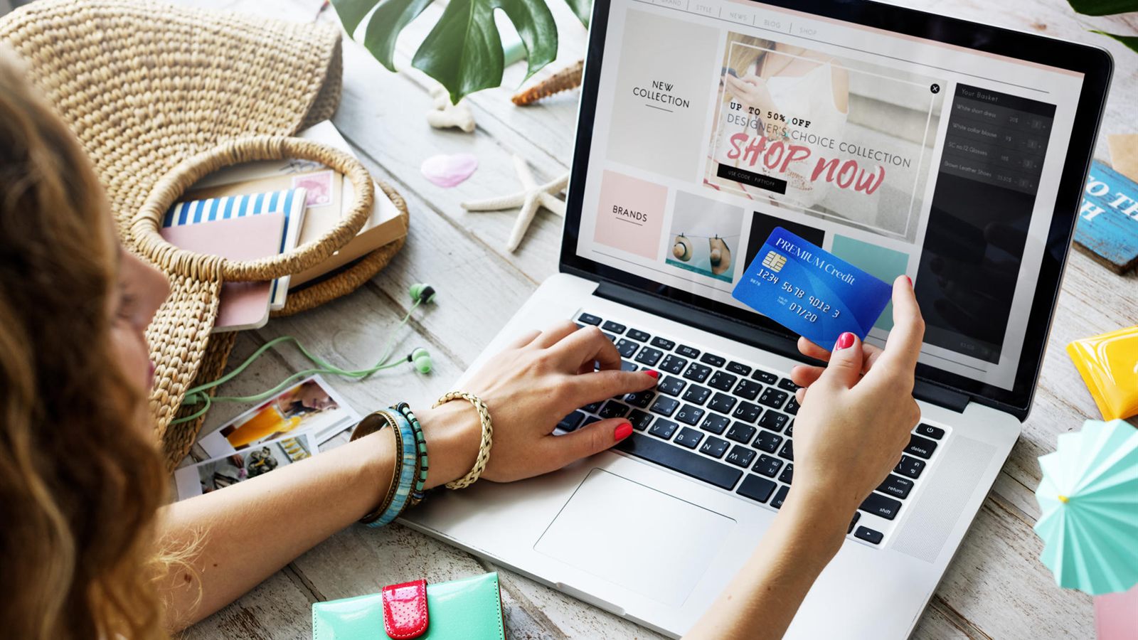 A woman shopping online on a laptop, holding a bank card, with a straw bag and a purse on the table beside her