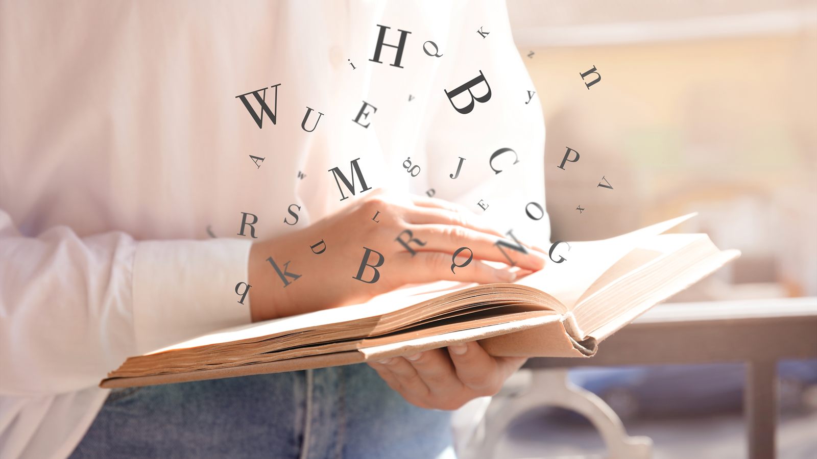 A person holding an open book, with printed letters of the alphabet floating up from its pages