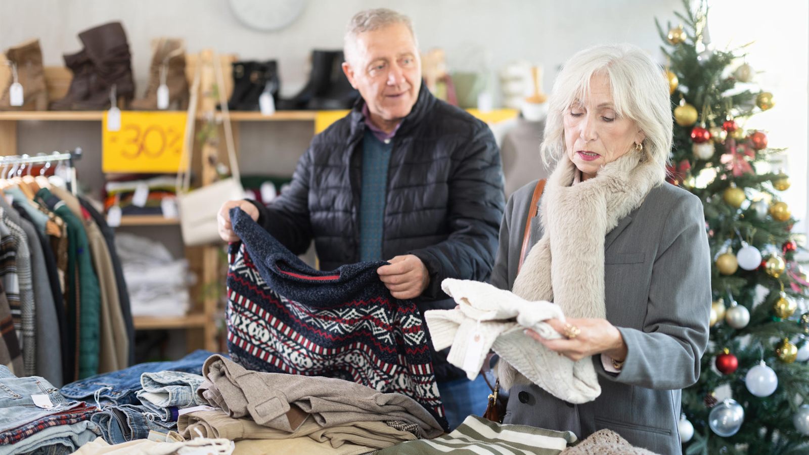 An older man and woman looking through second-hand jumpers on a table of folded clothes in a shop, with a decorated Christmas tree behind them